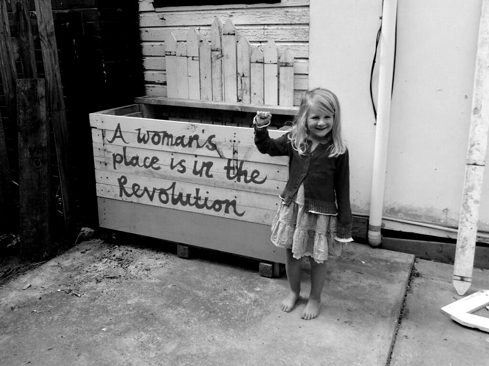 An incredibly cheery-looking little girl pumps her fist in front of some wood with the words "A woman's place is the revolution" painted on it.